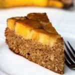 Fatia de bolo de banana invertido com garfo ao lado sobre prato branco em mesa branca, com bananas ao fundo