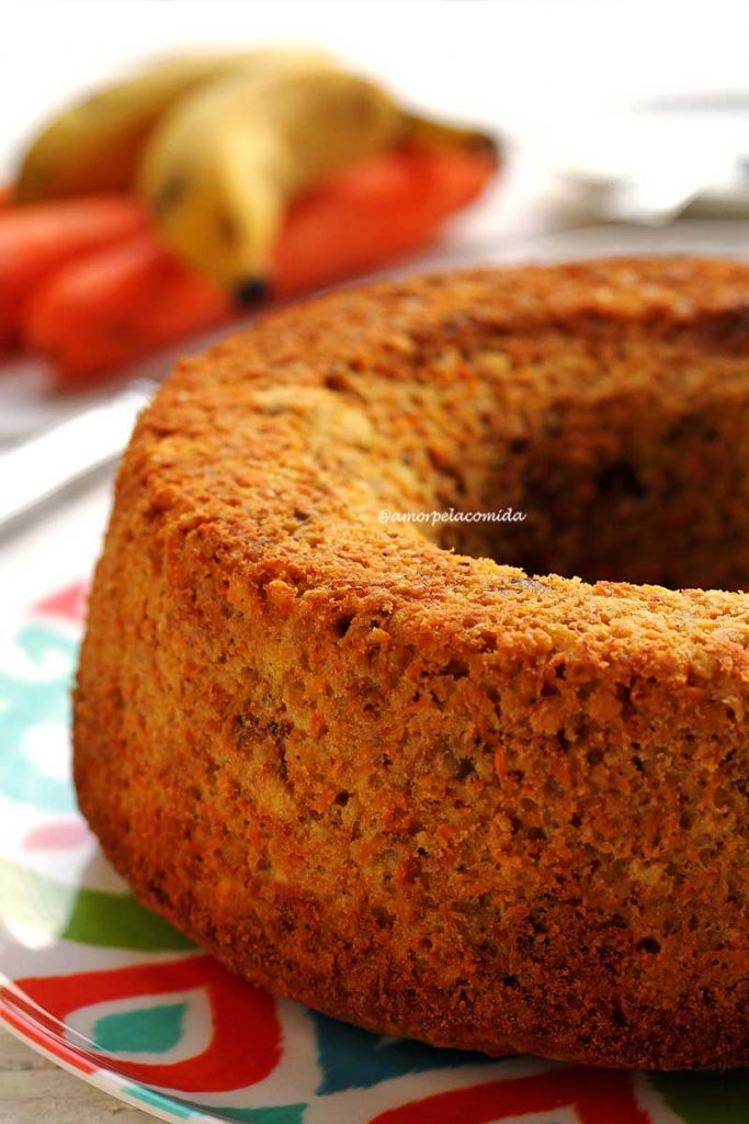 Bolo de banana com cenoura redondo com furo no meio sobre prato colorido em mesa branca