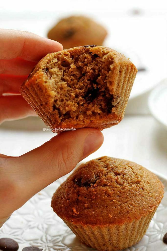 BOLO DE BAUNILHA FIT SEM GLÚTEN SEM LACTOSE COM RESÍDUO LEITE VEGETAL Mão segurando bolo de baunilha com gotas de chocolate pequeno mordido sobre mesa branca com outro bolinho pequeno sobre prato redondo branco