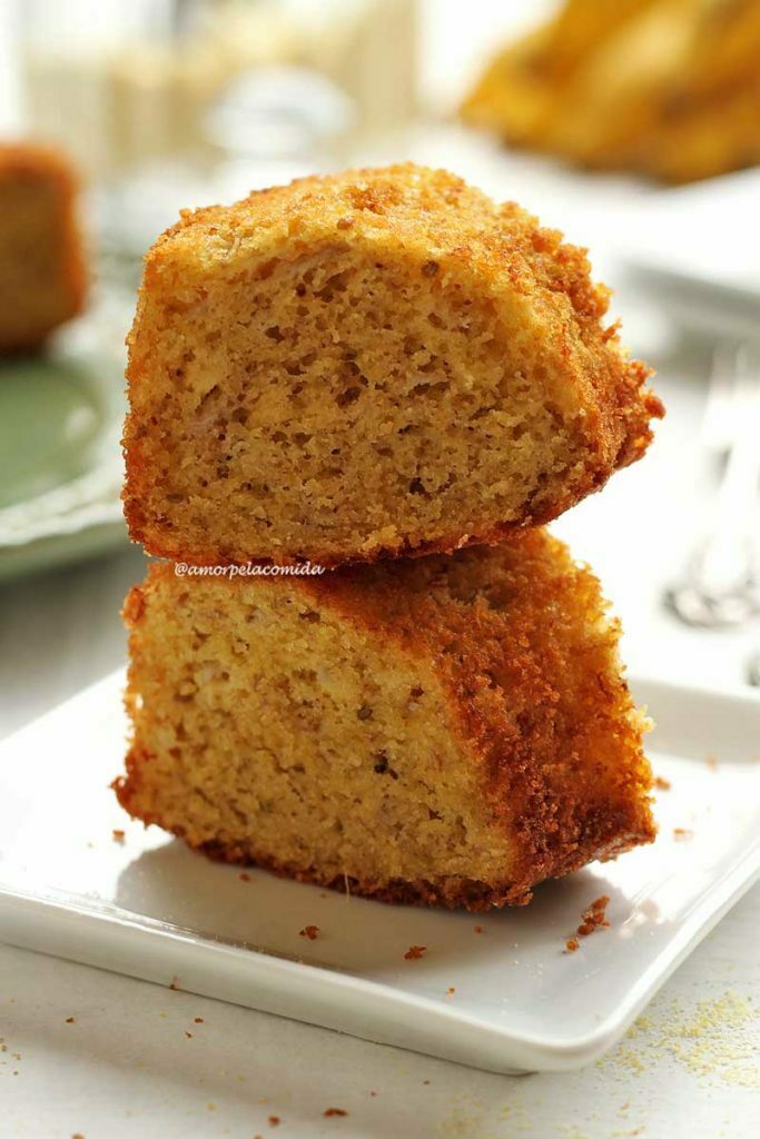 BOLO DE FUBÁ SIMPLES E RÁPIDO SEM GLÚTEN SEM LACTOSE Duas fatias de bolo de fubá sobrepostas em um prato branco quadrado pequeno em mesa branca