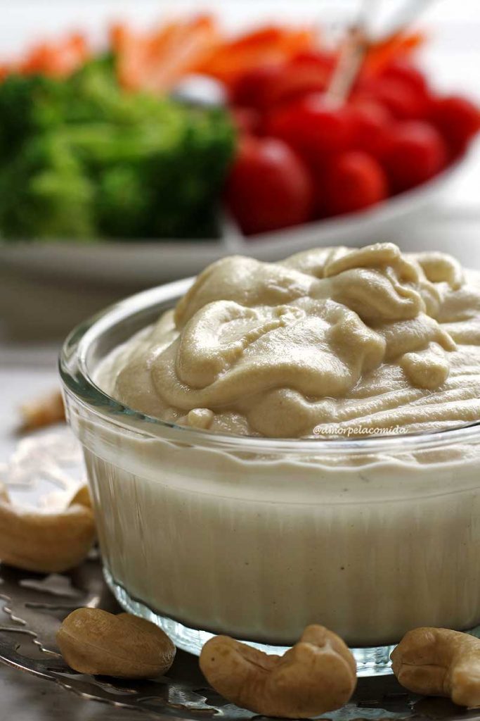 REQUEIJÃO CREMOSO CASEIRO FÁCIL VEGANO Pote de requeijão cremoso sobre um prato pequeno de prata com castanha de caju em volta e ao fundo vegetais em um prato branco