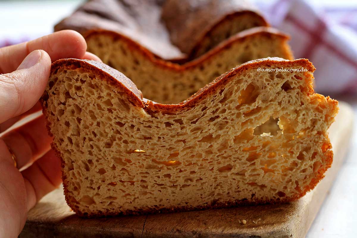 Pão de batata que deu errado e afundou no centro, mão segurando uma fatia e ao fundo o pão que deu errado