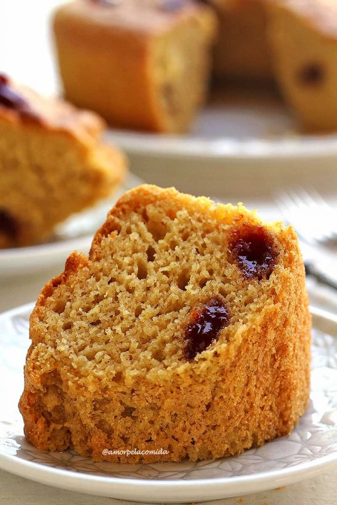 BOLO DE FUBÁ COM GOIABADA SEM GLÚTEN SEM LACTOSE FOFINHO Uma fatia de bolo de fubá com pedaços de goiabada sobre prato quadrado branco, ao fundo mais uma fatia cortada e o bolo desfocado
