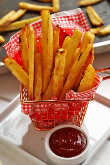 Cestinha de metal pequena com guardanapo vermelho com xadrez branco, dentro vários toletes de batata frita assada no forno, ao lado da cestinha um pote pequeno com ketchup, ao fundo a forma onde foram assadas as batatas