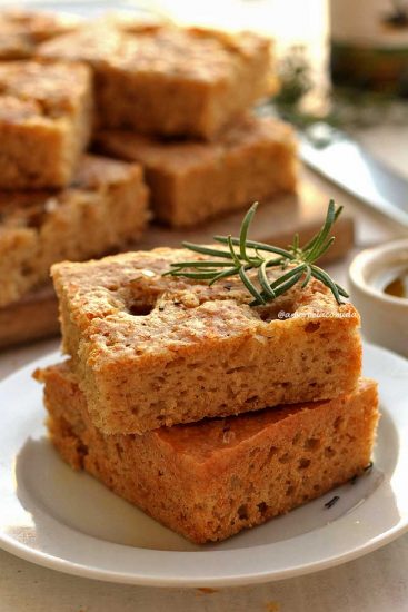Dois pedaços quadrados de focaccia sobre prato branco, ao fundo um pote de azeite pequeno e vários pedaços cortados de focaccia sobre uma tábua de madeira