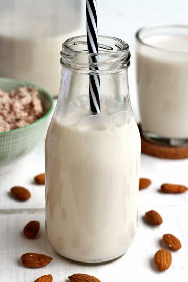 Garrafinha de vidro pequena com leite vegetal de amêndoas e um canudo preto e branco listrado, ao fundo mais leite dentro de vidros e o resíduo do leite. Na mesa branca algumas amêndoas espalhadas