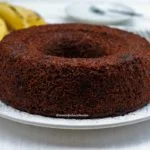 Bolo redondo com furo no meio de banana com chocolate sobre um prato branco, ao fundo bananas desfocadas e pratinhos pequenos desfocados