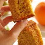 Mão segurando pedaço de bolo de laranja mordido mostrando a textura aerada da massa, abaixo uma fatia de bolo sobre um prato quadrado branco pequeno e laranjas desfocadas sobre uma mesa branca