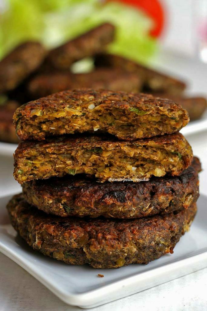 HAMBÚRGUER VEGETARIANO FÁCIL Dois hambúrgueres sobrepostos em um prato pequeno branco, sobre os hambúrgueres inteiros tem duas metade de um hambúrguer partido para mostrar a textura interna