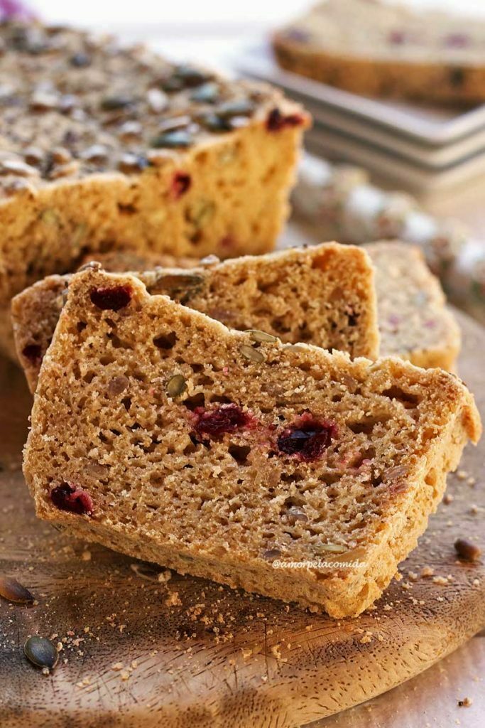 Três fatias de pão apoiadas umas nas outras em uma tábua de madeira, a fatia tem pedacinhos de cramberries e sementes dentro da massa