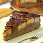 Fatia triangulas de bolo de banana com cobertura de banana caramelizada, ao fundo o bolo partido e algumas bananas em uma mesa branca