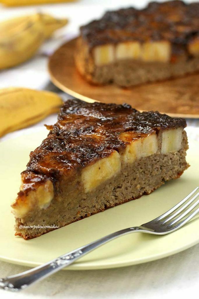 BOLO DE BANANA CARAMELIZADA SEM FARINHA COM AVEIA Fatia triangulas de bolo de banana com cobertura de banana caramelizada, ao fundo o bolo partido e algumas bananas em uma mesa branca