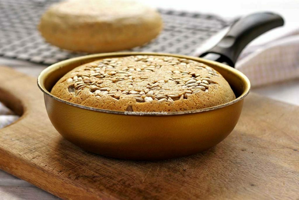 PÃO DE AVEIA NA FRIGIDEIRA Pão pequeno feito dentro de uma frigideira dourada, no topo do pão sementes de girassol. A frigideira está sobre uma tábua de madeira e ao fundo tem uma grelha preta com mais um pão redondo pequeno de frigideira desfocado da cena principal