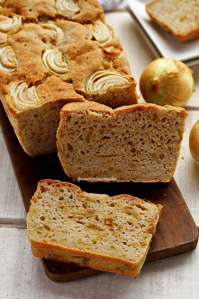Pão de cebola sobre tábua de madeira retangular escura, sobre o pão fatias de cebola, o pão está fatiado com 1 fatia apoiada no pão cortado e a outra fatia deitada na tábua