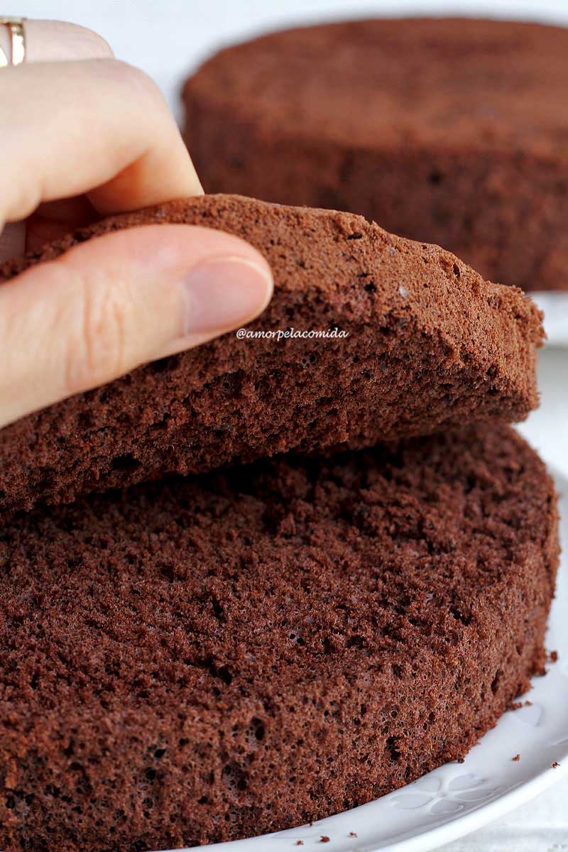 Mão levantando fatia de pão de ló de chocolate que foi cortado ao meio para mostrar a textura aerada interna da massa