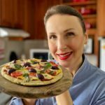 Mulher loira em uma cozinha com uniforme azul de chef segurando uma tábua de madeira, sobre a tábua a pizza de frigideira