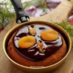 Frigideira pequena dourada sobre tábua de madeira, dentro um panetone decorado com cobertura de chocolate cremosa com damasco e nozes