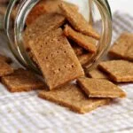 Vários biscoitinhos saindo de um vidro transparente apoiados sobre pano quadriculado branco e bege