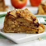 Fatia de torta de maçã sobre prato branco pequeno mostrando todas as camadas de maçã e algumas passas, no topo flocos de aveia