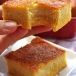 Mão segurando pedaço de bolo de fubá com laranja mordido, abaixo mais uma fatia sobre prato quadrado pequeno, ao fundo uma forma vermelha sobre uma mesa branca