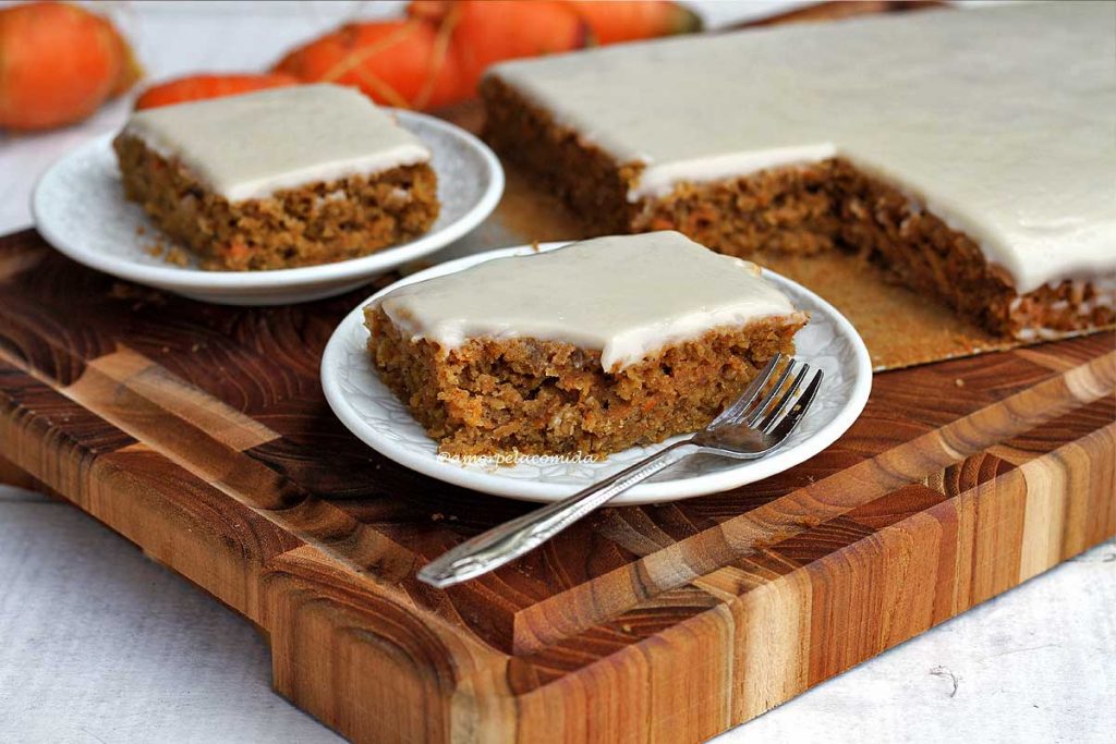 Dois pedaços de bolo de cenoura com cobertura branca sobre uma tábua de madeira, ao lado o restante do bolo, ao fundo algumas cenoura desfocadas