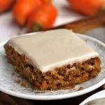 Pedaço de bolo de cenoura com cobertura branca em um prato pequeno redondo, ao fundo algumas cenouras desfocadas