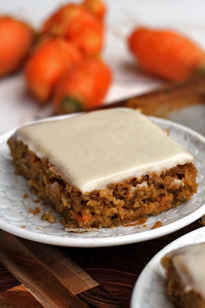 Pedaço de bolo de cenoura com cobertura branca em um prato pequeno redondo, ao fundo algumas cenouras desfocadas