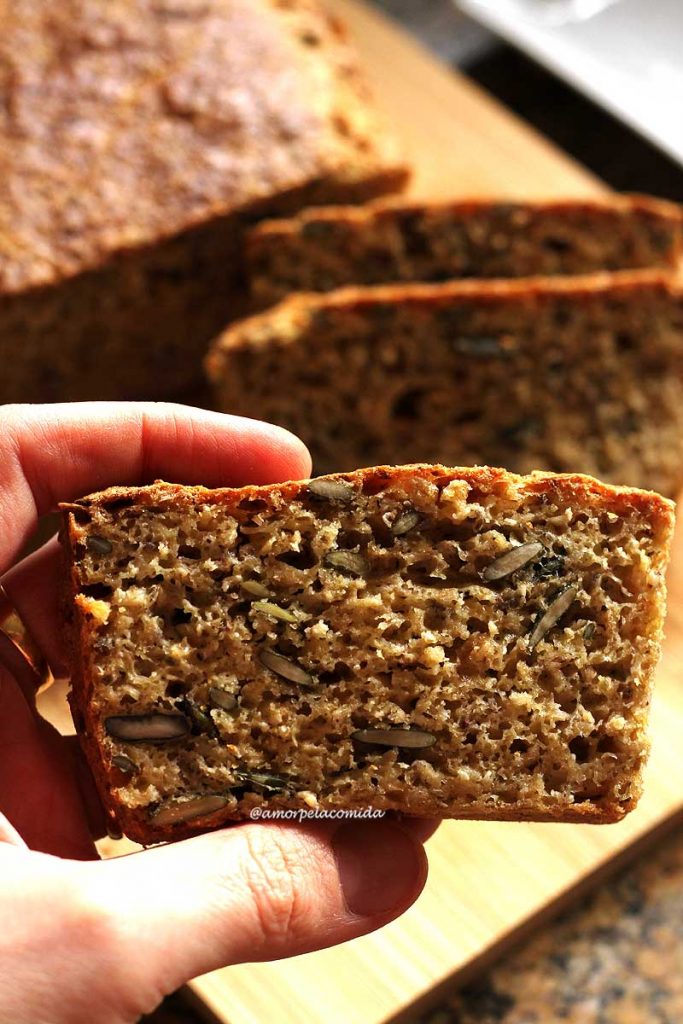 Pão segurando fatia de pão de grão-de-bico, é possível visualizar várias sementes na fatia, ao fundo mais fatias de pão e o pão caseiro desfocado
