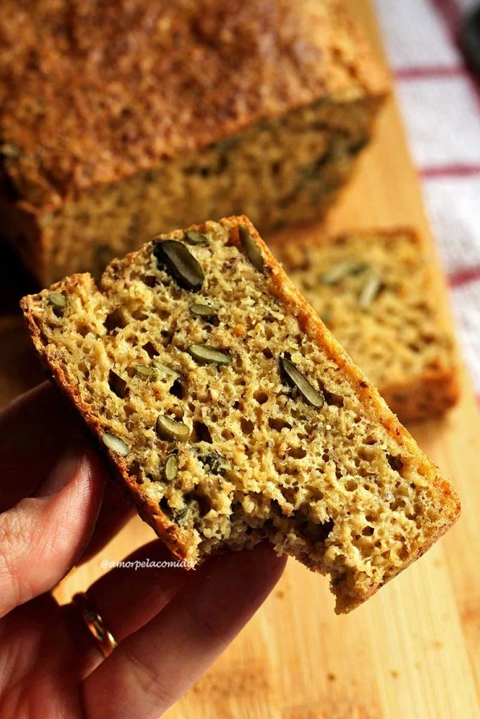 Mão segurando fatia de pão caseiro mordido, abaixo o restante do pão de forma e uma fatia sobreposta em uma tábua de bambu