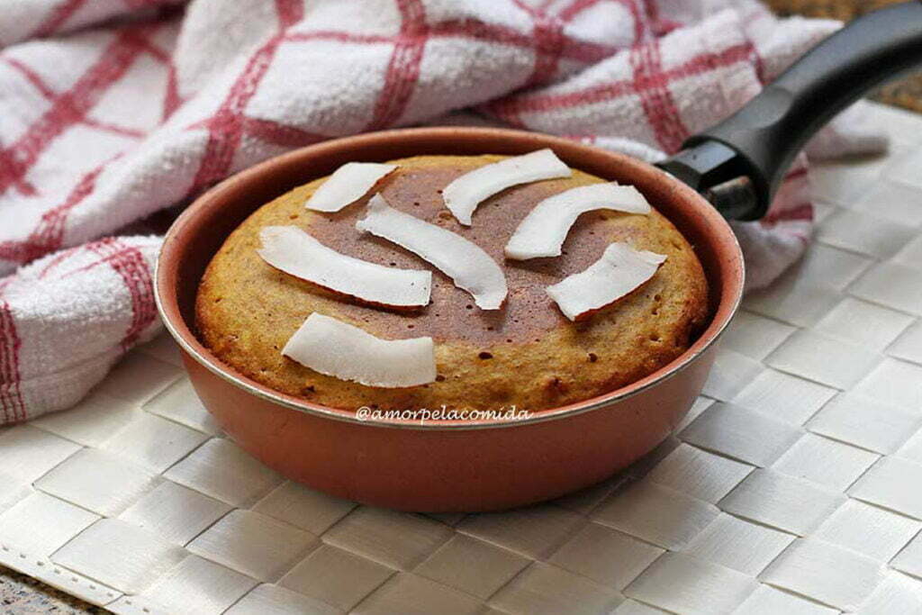 Frigideira pequena com bolo de abóbora com coco dentro, sobre o bolo lâminas de coco fresco, a frigideira está sobre um jogo americano branco, ao fundo uma toalha branca e vermelha quadriculada