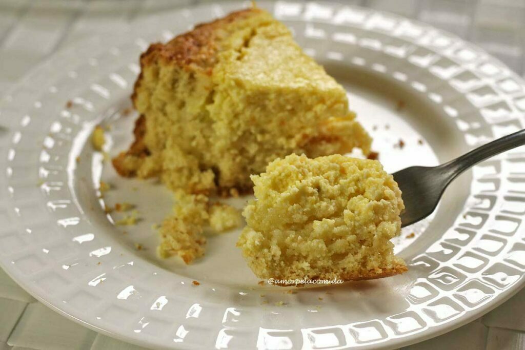 Fatia de bolo de queijo sobre prato redondo branco, tem um garfo tirando um pedaço da fatia, mostrando a parte cremosinha e granulada do queijinho e na parte inferior o bolo
