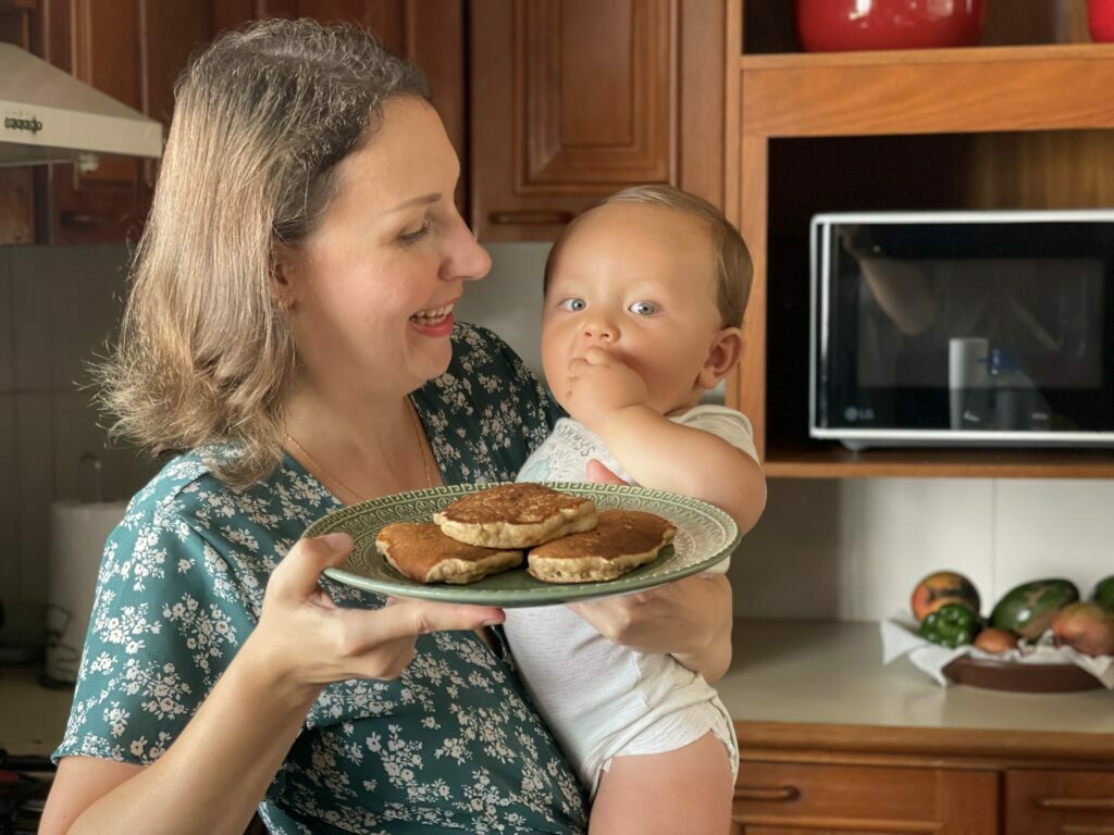 Mulher loira segurando criança no coloco, a criança está comendo um pedaço de panqueca que pegou do prato que a mãe está segurando