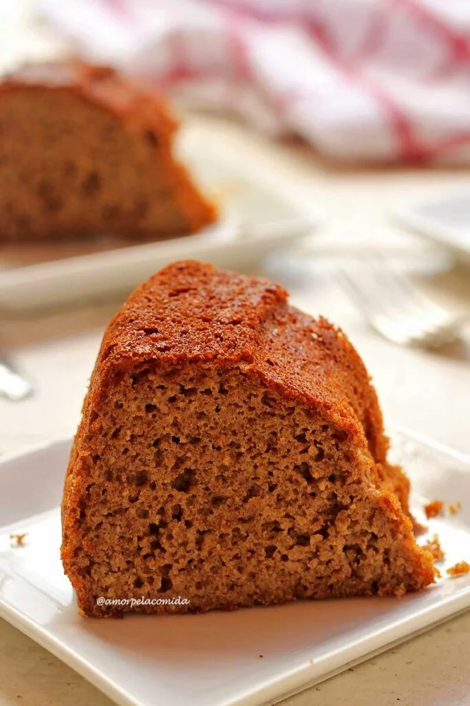 Pedaço de bolo sobre prato quadrado pequeno sobre mesa branca, ao fundo aparece uma parte de outra fatia de bolo sobre o prato