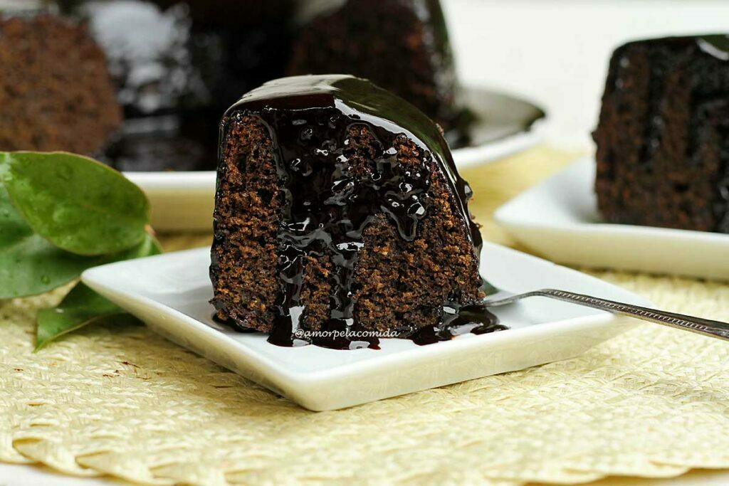 Fatia de bolo sobre prato quadrado branco, o bolo é de chocolate com cobertura de chocolate que está no topo da fatia e escorre pelas laterais