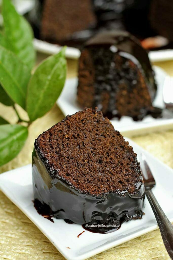 Duas fatias de bolo de chocolate com cobertura de chocolate, a da frente está deitada sobre o prato e a de trás está de pé, ao lado um galho de ora-pro-nóbis