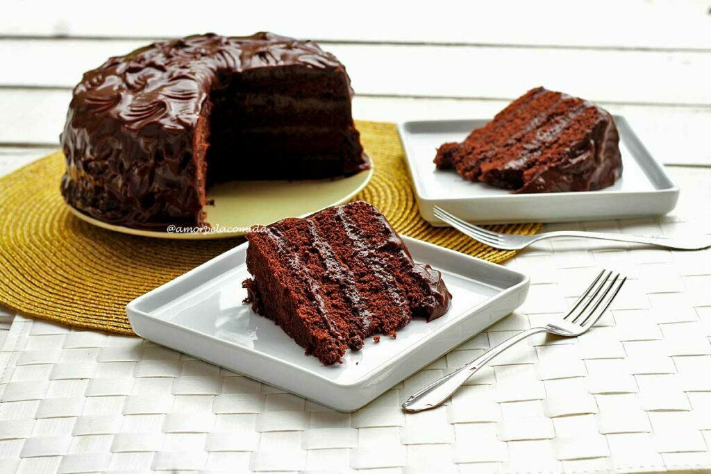 Bolo de chocolate recheado com recheio e cobertura de chocolate, ao lado do bolo partido estão duas fatias de bolo sobre 2 pratos de bolo quadrados