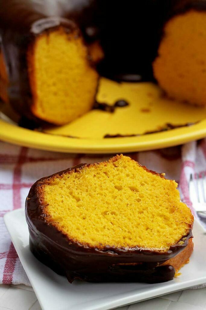 Fatia de bolo de cenoura deitada sobre prato quadrado pequeno, ao fundo o bolo partido sobre um prato amarelo