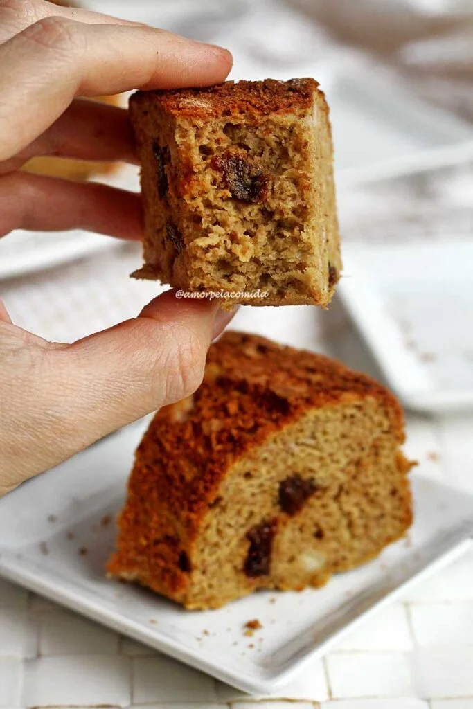 Mão segurando fatia de bolo mordido, abaixo uma fatia inteira sobre um prato quadrado pequeno