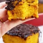 Mão segurando pedaço de bolo de cenoura com cobertura de chocolate que já foi mordido, abaixo mais um pedaço sobre prato quadrado pequeno