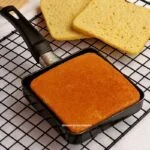 Frigideira preta quadrada sobre grade, dentro da frigideira um pão e atrás mais um pão quadrado partido ao meio