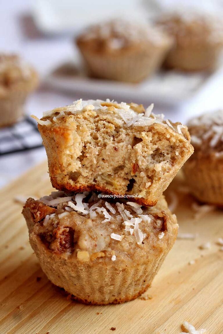 BOLO DE BANANA COM AVEIA INTEGRAL SEM AÇÚCAR Bolinho de banana com cobertura de coco em flocos mordido sobre outro bolinho inteiro em uma tábua de madeira