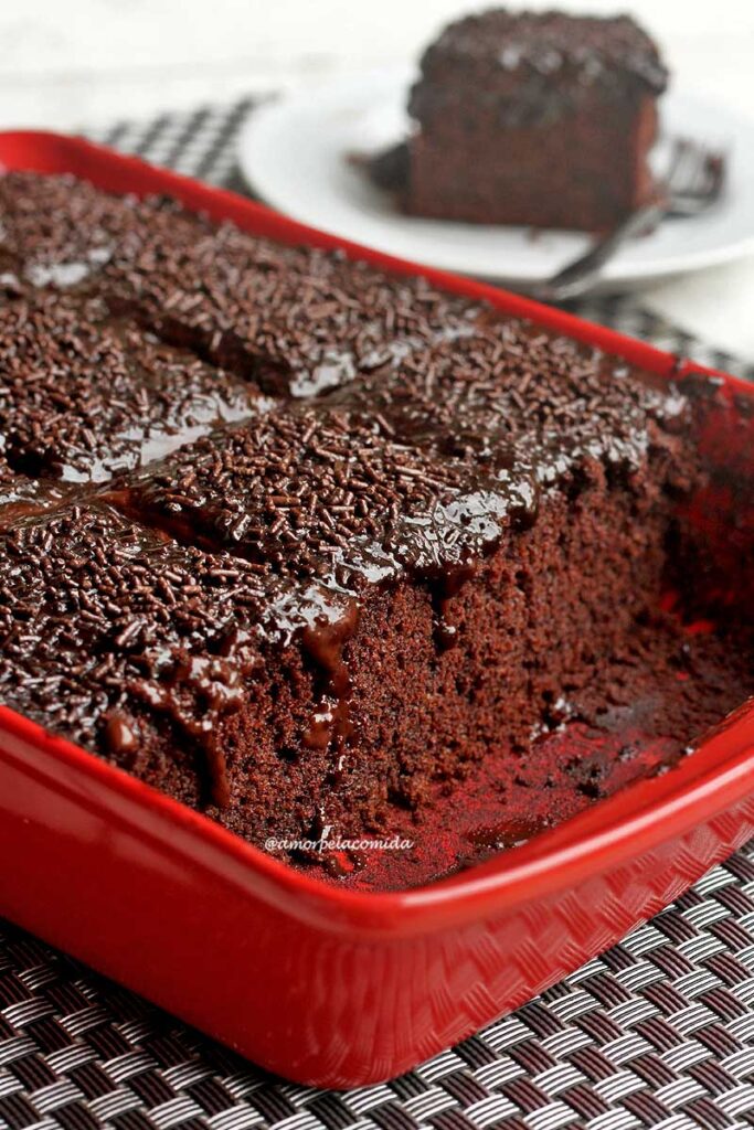 Forma retangular vermelha com bolo de chocolate dentro, o bolo tem cobertura cremosa de chocolate e granulado por cima