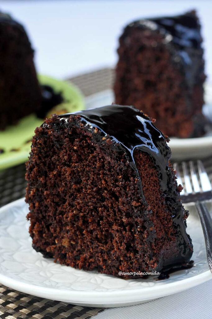 Fatia de bolo de chocolate com cobertura de chocolate sobre prato redondo pequeno, ao lado um garfinho