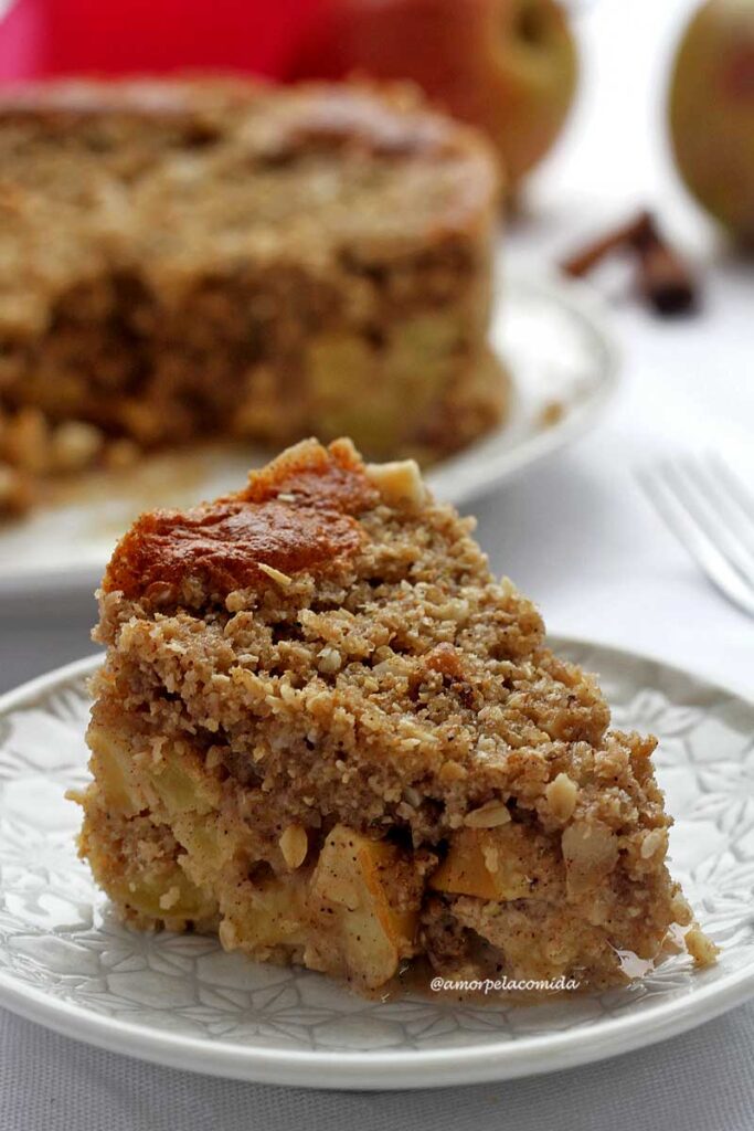 Fatia de torta de maçã com cobertura de farofa crocante sobre prato redondo, ao fundo a torta de maçã partida