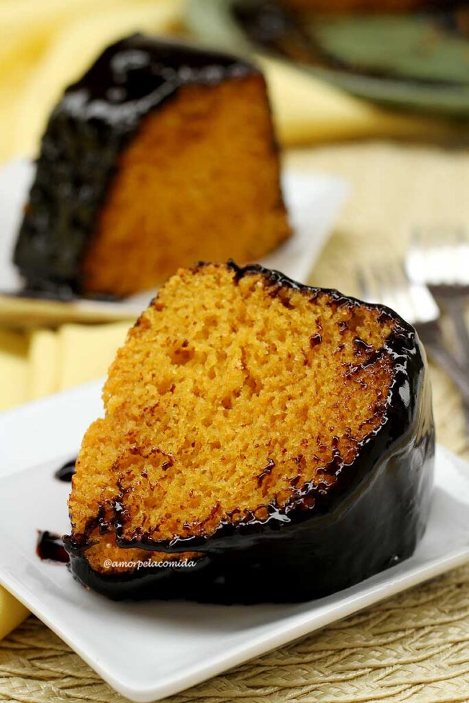 Uma fatia de bolo de cenoura com cobertura de chocolate deitada sobre prato quadrado pequeno, ao fundo mais uma fatia em outro prato