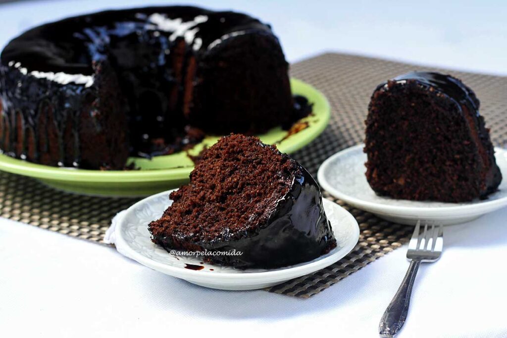 Dois pedaços de bolo de chocolate sobre prato redondo pequeno ao fundo o bolo partido