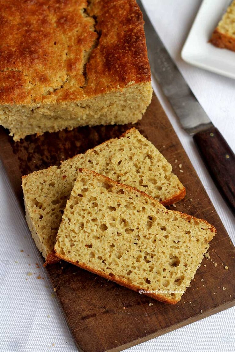 Duas fatias de pão sobrepostas sobre uma tábua de madeira retangular de cor escura, ao lado uma faca e o pão que foi cortado as fatias