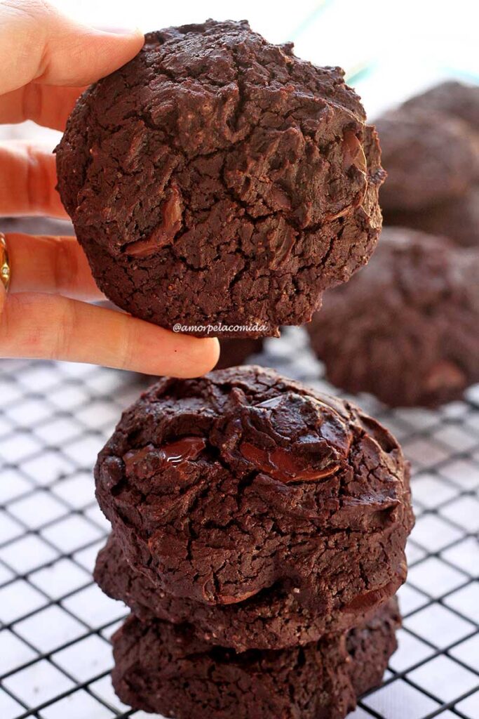 Mão segurando biscoito grande de chocolate, abaixo uma torre de biscoito sobre uma grade preta