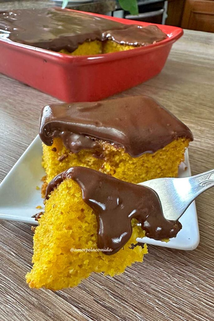 Pedaço de bolo de cenoura com cobertura de chocolate sobre um garfo pequeno, ao fundo o pedaço partido sobre um prato quadrado pequeno