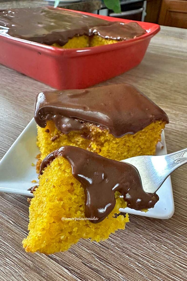 RECEITA DE BOLO DE CENOURA FOFINHO DE LIQUIDIFICADOR COM AVEIA Pedaço de bolo de cenoura com cobertura de chocolate sobre um garfo pequeno, ao fundo o pedaço partido sobre um prato quadrado pequeno